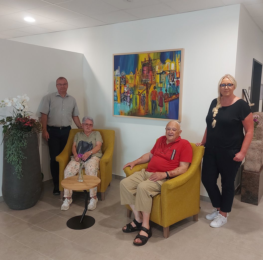 Zwei ältere Personen sitzen in gelben Sesseln, zwei weitere Personen stehen daneben in einem modernen Raum mit großem grauem Blumentopf und farbenfrohem Bild an der Wand.