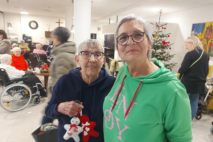 Zwei ältere Personen stehen in einem Raum mit Weihnachtsbaum, im Hintergrund sitzen weitere Menschen, darunter eine Person im Rollstuhl.