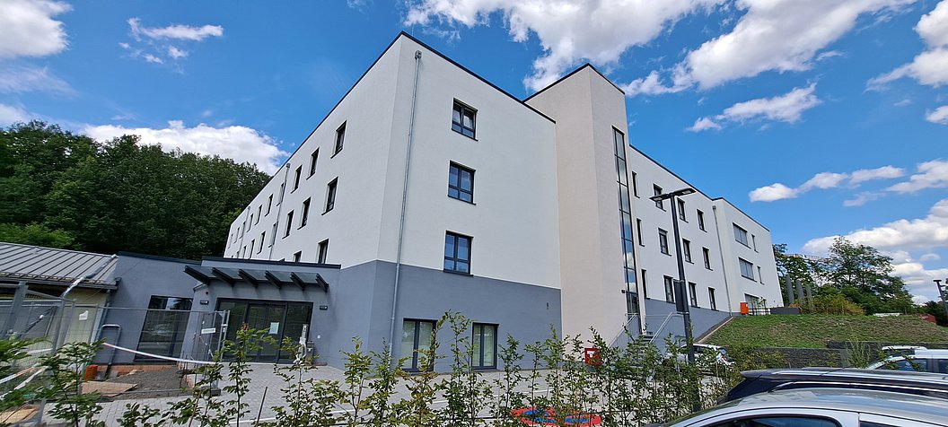 Mehrstöckiges, weiß-graues Wohngebäude mit mehreren Fenstern unter blauem Himmel mit Wolken.