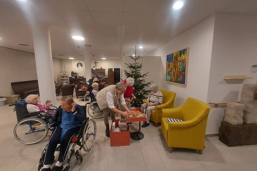 Mehrere ältere Menschen, einige im Rollstuhl, sitzen in einem Gemeinschaftsraum mit Weihnachtsbaum, während eine Person an einem kleinen Tisch etwas arrangiert.