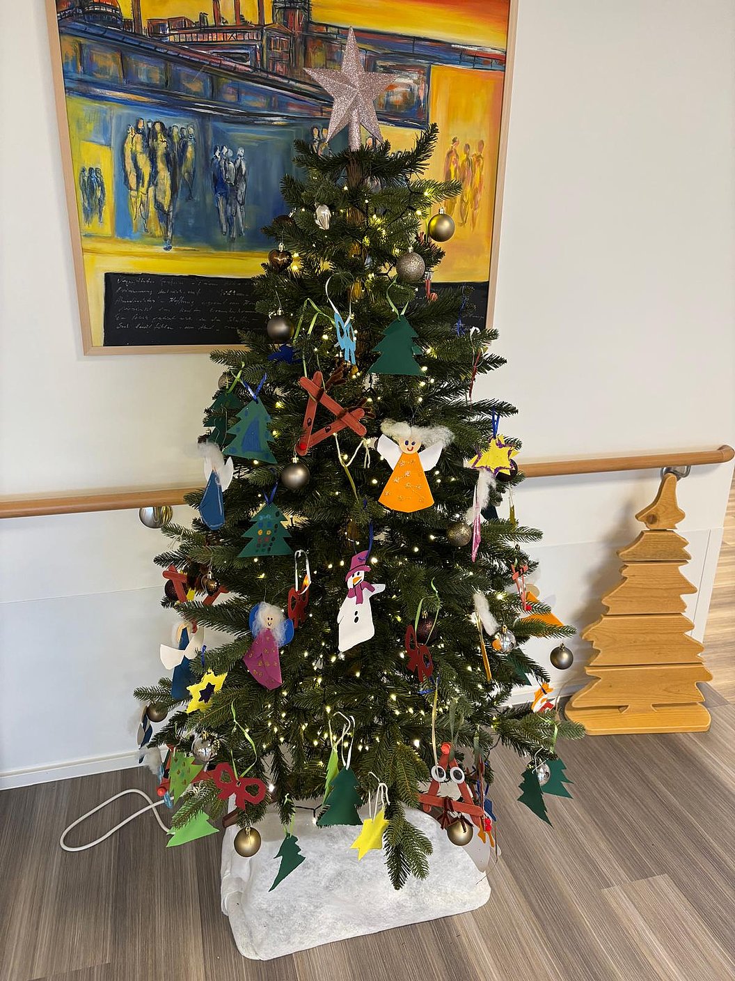Weihnachtsbaum mit selbstgebasteltem Schmuck aus Filz und Papier, darunter Engel, Sterne und Tannenbäume, vor einem Gemälde und neben einer Holzbaum-Dekoration.
