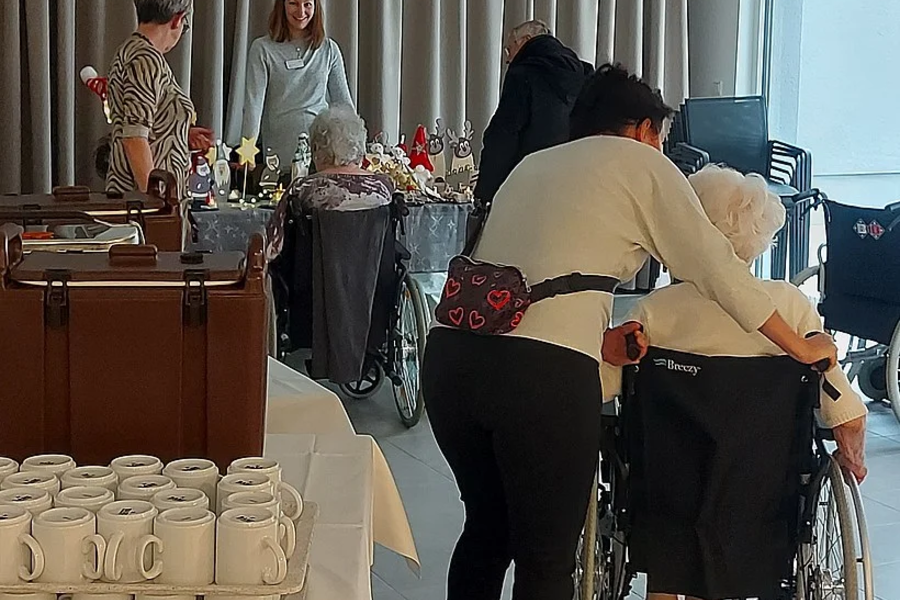 Mehrere Personen in einem Raum, eine Person stützt eine ältere Person im Rollstuhl, im Vordergrund ein Tisch mit gestapelten weißen Tassen.
