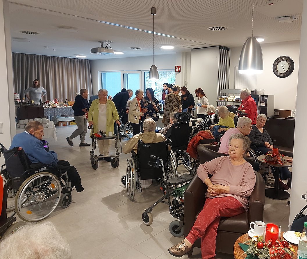 Mehrere ältere Menschen, einige im Rollstuhl, versammeln sich in einem Gemeinschaftsraum mit Sitzgelegenheiten und einem Buffet im Hintergrund.