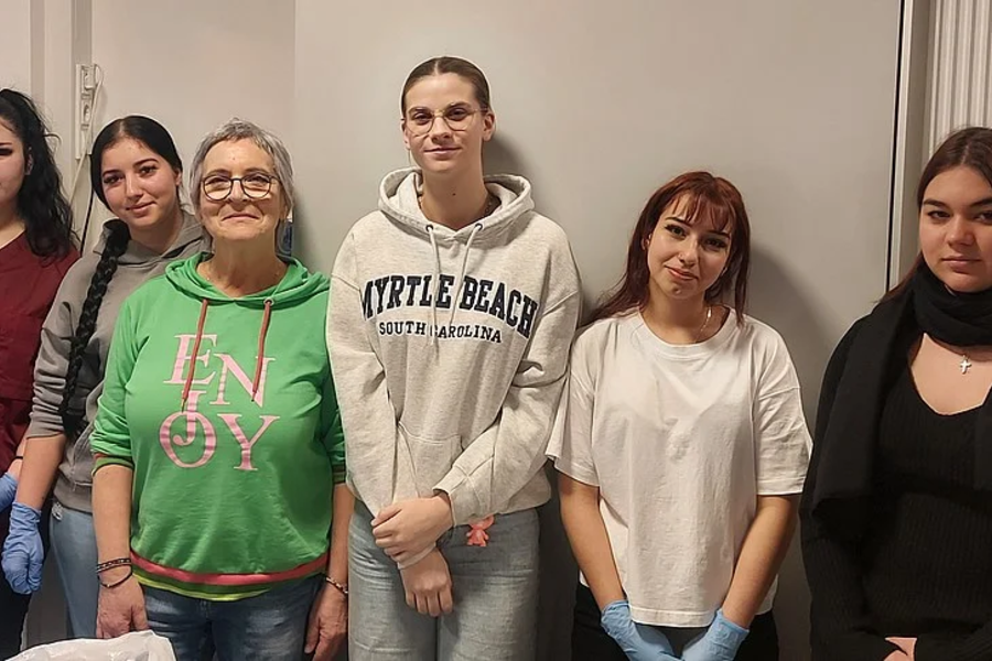 Sechs Personen stehen nebeneinander vor einer Wand, alle mit unterschiedlichen Oberteilen und entspannten Haltungen.