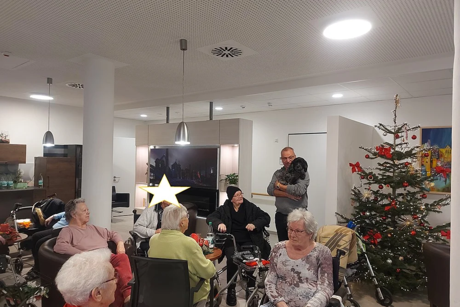Mehrere ältere Menschen sitzen in einem Wohnzimmer mit Weihnachtsbaum und Fernseher, eine Person hält ein Kind im Arm.
