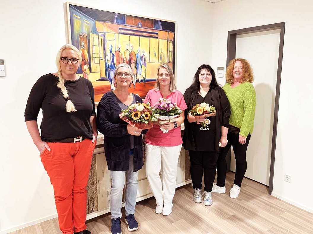 Fünf Frauen stehen in einem Raum mit Holzfußboden vor einer farbigen Wandmalerei, drei von ihnen halten Blumensträuße.