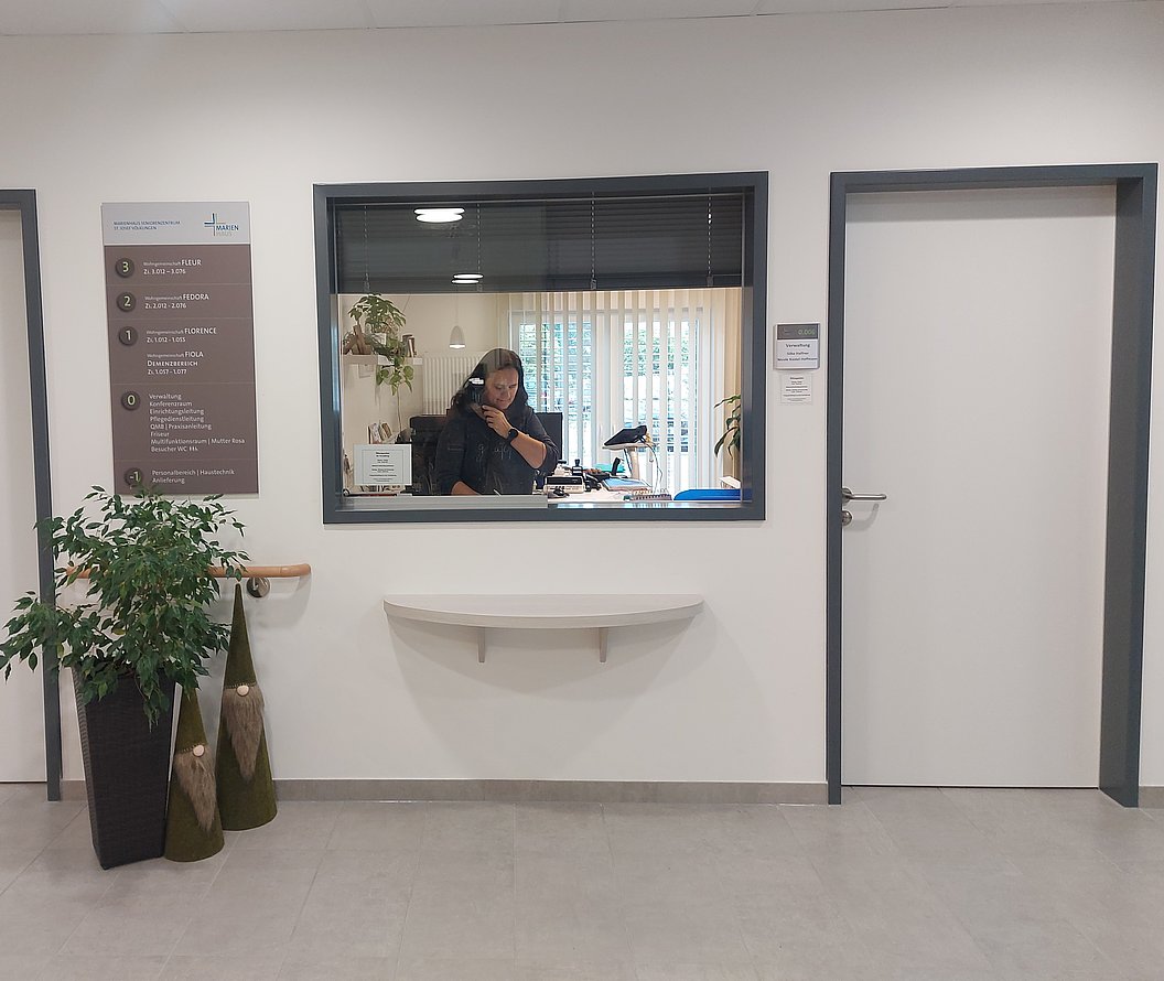 Empfangstresen mit Fenster in einer hellen, modernen Innenraumumgebung, links daneben eine Pflanze und ein Informationsschild an der Wand.