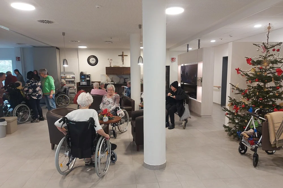 Mehrere ältere Menschen sitzen und stehen in einem hellen Gemeinschaftsraum mit Weihnachtsbaum und Säule in der Mitte.