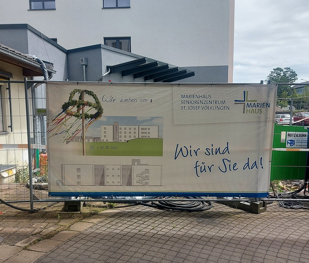 Bauzaun mit Banner des Marienhauses Seniorenzentrums St. Josef Völklingen, das Umzug ankündigt, mit schematischer Gebäudezeichnung und Text 'Wir ziehen um' sowie 'Wir sind für Sie da!'.