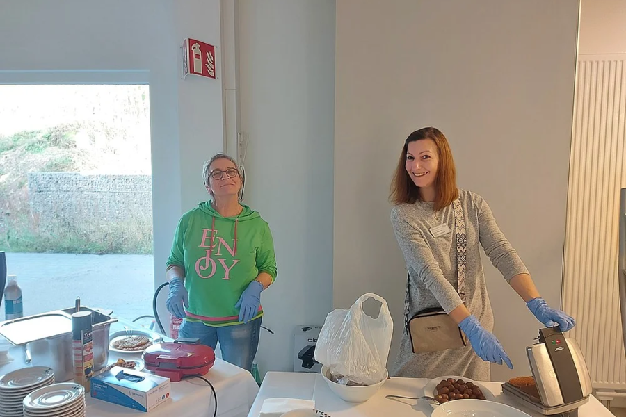 Zwei Personen mit blauen Handschuhen bereiten an einem Tisch mit Tellern, einer Waffelmaschine und Lebensmitteln Speisen zu.