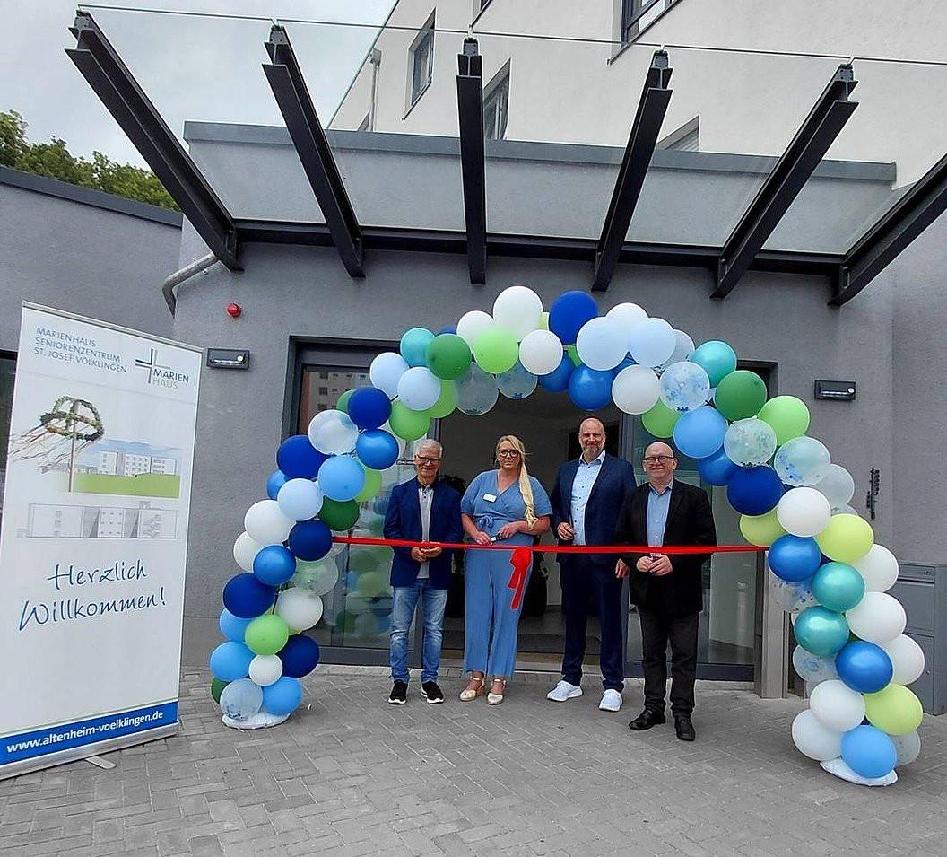Vier Personen stehen vor einem Gebäude unter einem Bogen aus weißen, blauen und grünen Luftballons und halten ein rotes Band