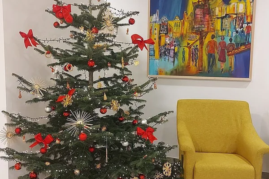 Weihnachtsbaum mit roten Schleifen und Kugeln neben gelbem Sessel vor weißer Wand mit farbenfrohem Gemälde.