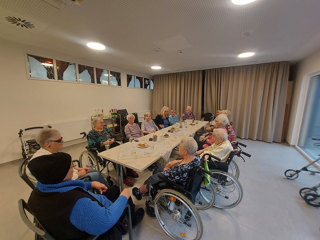 Mehrere ältere Frauen sitzen in einem hellen Raum an einem langen Tisch, einige im Rollstuhl, andere auf Stühlen, mit Getränken und kleinen Snacks auf dem Tisch.