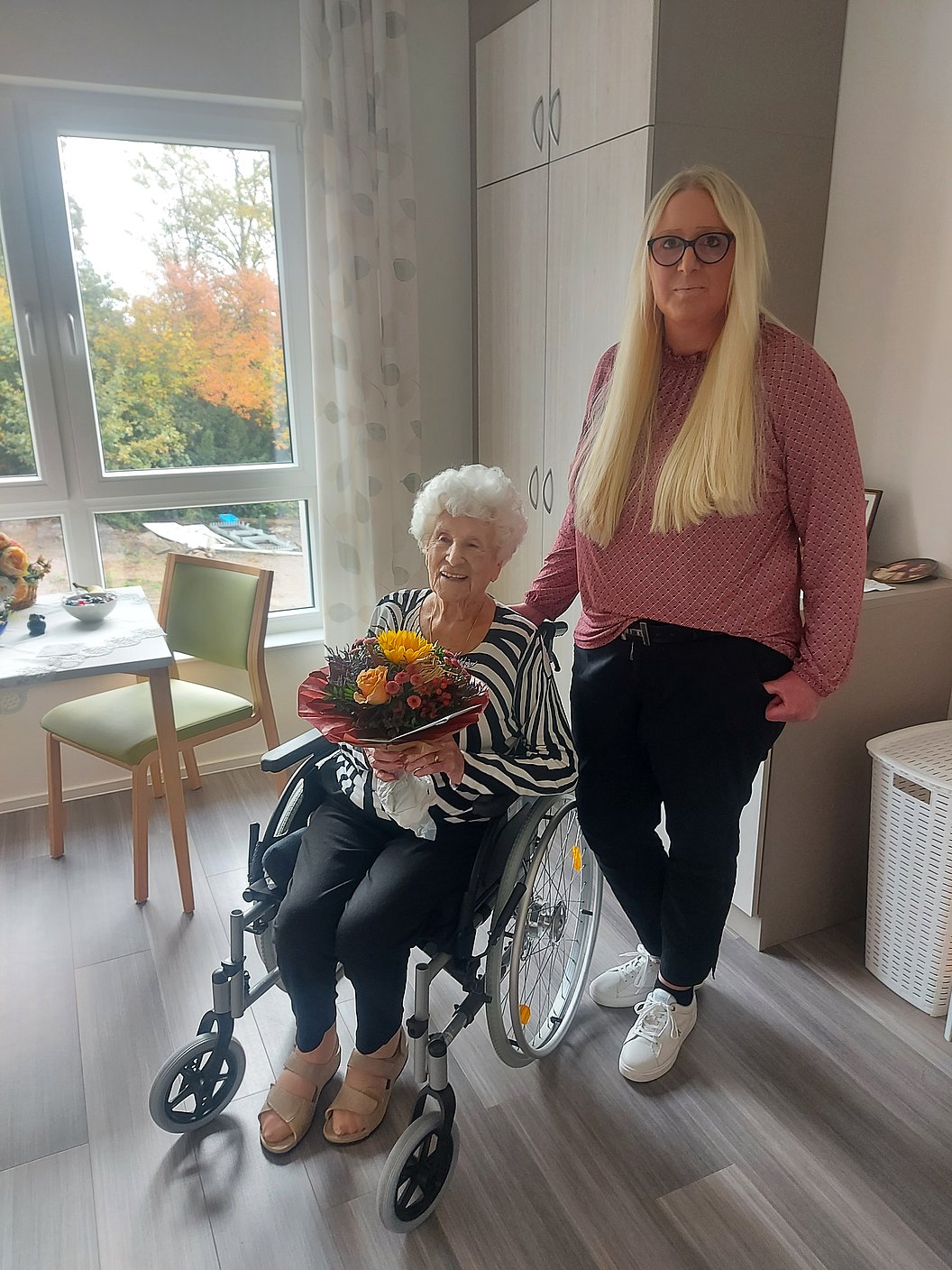 Ältere Person im Rollstuhl hält einen Blumenstrauß, neben ihr steht eine jüngere Frau mit langen blonden Haaren in einem roten Pullover und schwarzen Hosen in einem hellen Raum mit Fenster und Möbeln.