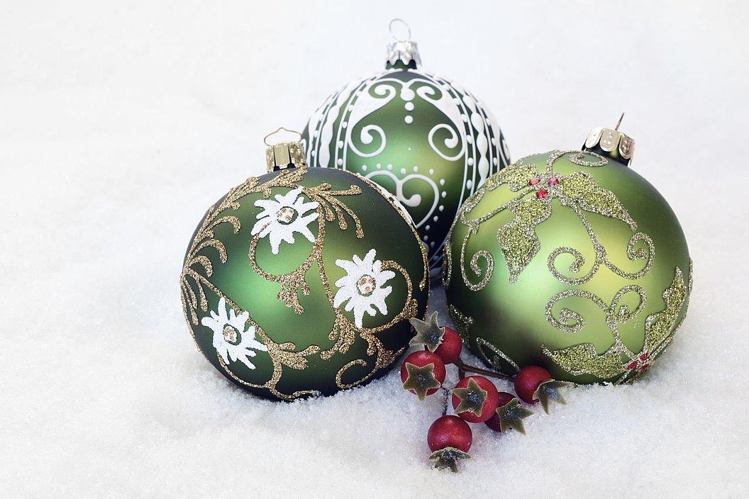 Drei grüne Christbaumkugeln mit goldenen und weißen Verzierungen liegen auf Schnee neben roten Beerenzweigen.
