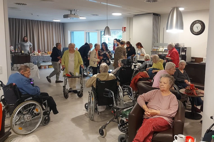 Mehrere ältere Menschen, einige im Rollstuhl oder mit Rollator, sitzen und stehen in einem Gemeinschaftsraum mit Tischen, Stühlen und einer Kaffeestation.