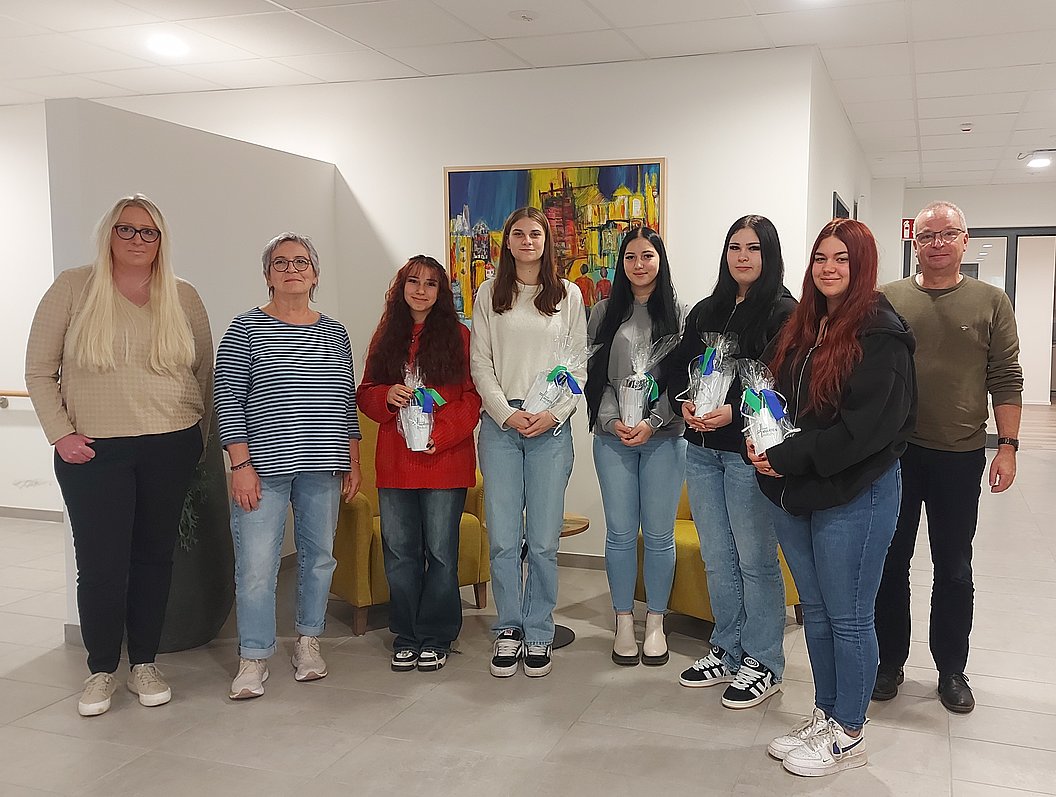 Gruppe von acht Personen steht in einem Raum, einige halten kleine Geschenke in Geschenkpapier mit blauer Schleife.