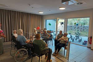 Gruppe älterer Menschen sitzt in einem Raum mit großen Fenstern und spielt mit einem Netz und einem Ball