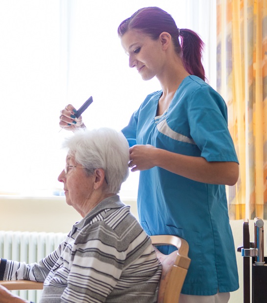 Pflegekraft in blauer Arbeitskleidung frisiert sitzende ältere Person mit grauen Haaren in einem hellen Raum